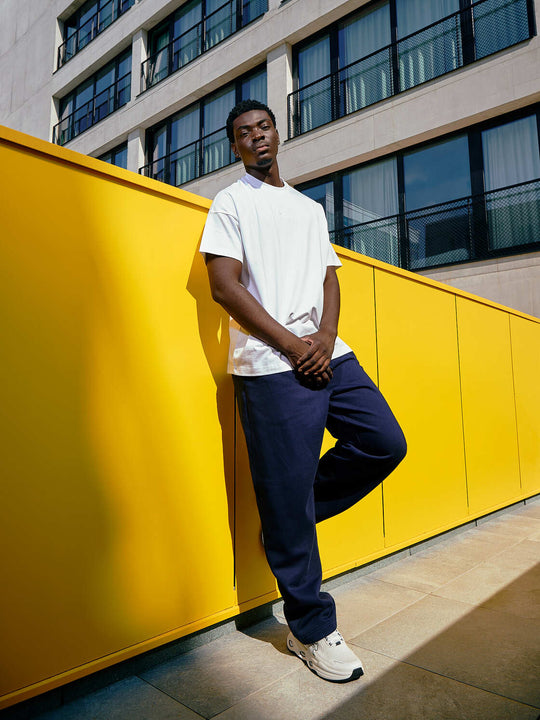 Un homme adossé à un mur orange porte un t shirt Airness blanc, un pantalon bleu et des baskets Namib blanc &amp; noir de chez Airness