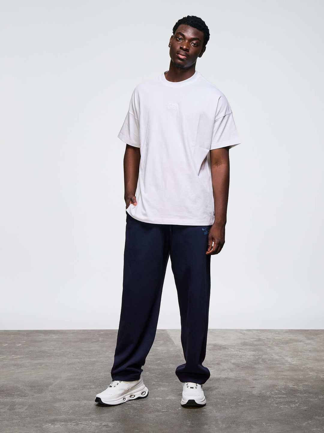 Un homme de face porte un t-shirt blanc modèle fabricio, un pantalon de jogging noir et des sneakers Namib Airness blanc &amp; noir