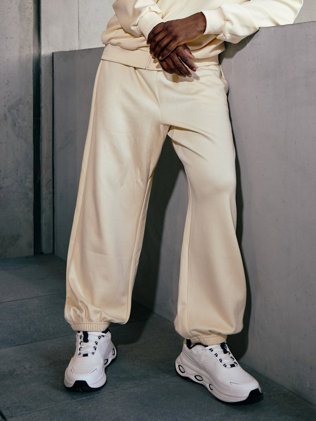 pantalon Evan Airness écru porté par un homme en survêtement et sneakers Airness accoudé à un muret en béton